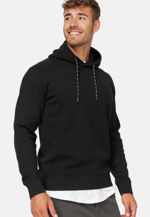 Sweatshirt à capuche noire en tricot texturé, avec un cordon de serrage, des poignets et un ourlet côtelés. Porté sur une chemise blanche, avec une coupe ajustée.