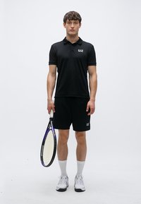 Juodas polo marškinėlis su EA7 logotipu, juodi šortai, balti sportiniai kojinės ir balti sportbačiai, laikantis violetinį teniso raketę.