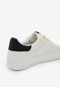 Sneaker bianco con un accento in suede nero sul tallone, lacci gialli, materiale in pelle liscia e una spessa suola in gomma bianca.