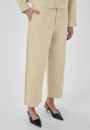 Béžové široké nohavice v cropped strihu s bočnými vreckami, nosené s čiernymi slingback lodičkami s špicatou špičkou na svetlom pozadí.