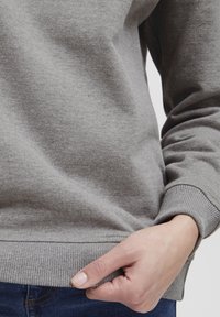 Felpa grigia realizzata in un morbido misto cotone, con polsini e orlo a costine, caratterizzata da una texture liscia e un design aderente. Mani visibili che tengono il fondo.
