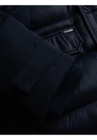 Manga de chaqueta acolchada azul oscuro con puño cosido y detalle de tira de Velcro, reposando sobre parte del bolsillo de la chaqueta con etiqueta metálica.