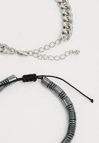 Sølv kædedesign med sammenkædede led, der inkluderer en sort snor, med cylindrisk metalperler og en knude-detalje.