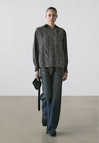 Gray knit vest over a dark shirt, paired with wide-leg dark jeans. The model holds a small black bag and wears black shoes. Simple background.