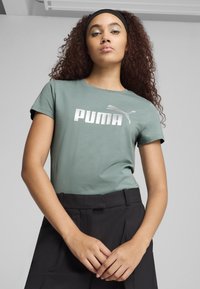 Zelené tričko s krátkým rukávem s velkým stříbrným logem "PUMA" a emblémem kočky, spárované s černými kalhotami s vysokým pasem. Hladká bavlněná textura.