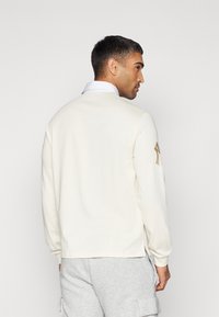 Long-sleeve cream sweatshirt with a small logo on the left sleeve, featuring a collared design and side slits at the hem.