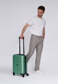 Hombre con camiseta blanca y pantalones grises de pie, con una mano en el bolsillo y sosteniendo una maleta verde con el asa extendida.
