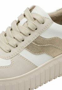 Baskets beige et blanches avec une semelle épaisse et texturée, des lacets en tissu beige et un motif métallique ondulé sur le panneau latéral.