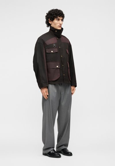 Άνδρας που φοράει μαύρο και σκουρόμωβ σακάκι εργασίας με πολλές τσέπες και γκρι παντελόνι φαρδιάς γραμμής, στέκεται μπροστά σε απλό φόντο.