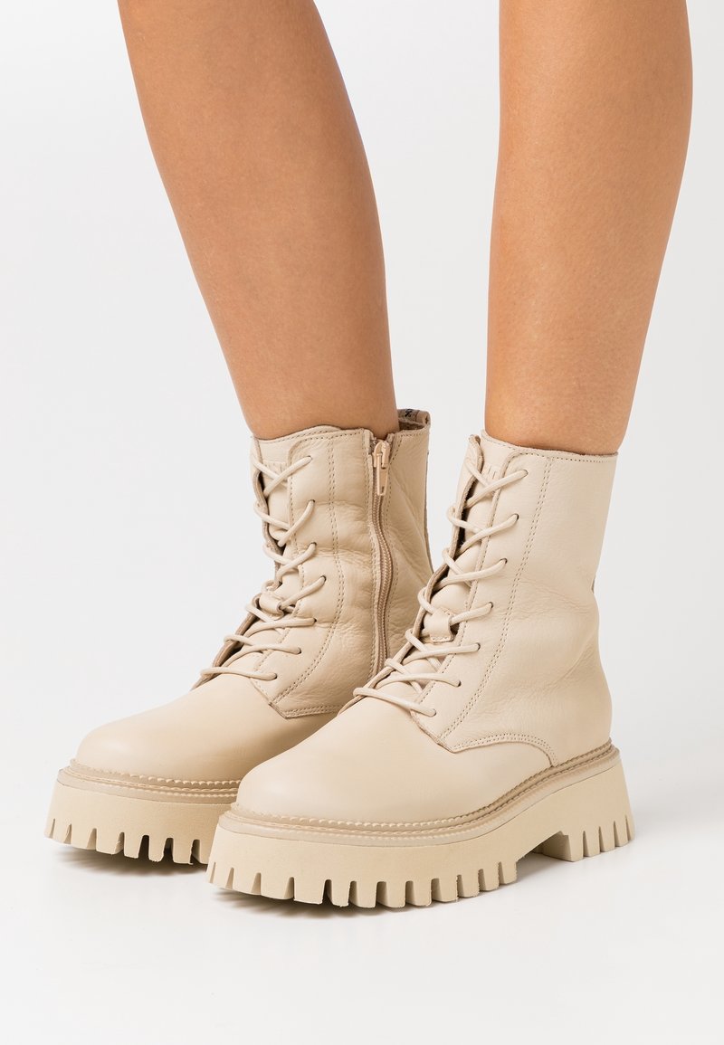 Jambes portant des bottines beiges à lacets avec semelles épaisses et résistantes et fermeture éclair latérale sur fond blanc.