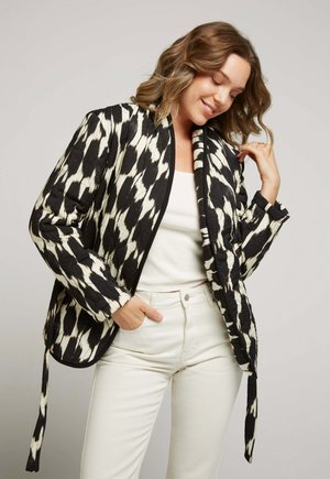 Veste matelassée à motifs noir et blanc avec un col châle, des manches longues et des liens à la taille, portée sur un haut blanc et un jean clair.