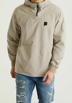 Homme portant une veste beige à capuche avec demi-fermeture éclair et poche avant, et un jean bleu déchiré, debout devant un fond clair uni.