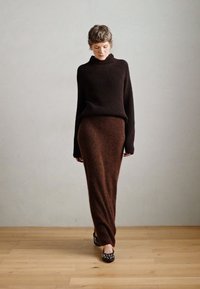 Maglione marrone lavorato a maglia con colletto alto, abbinato a una lunga gonna marrone. L'outfit presenta una morbida texture e una silhouette aderente. Scarpe nere con decorazioni.