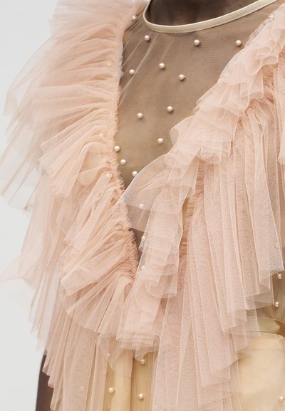 Φόρεμα από διάφανο μπεζ ύφασμα με μικρές πέρλες και μεγάλα στρωματοποιημένα απαλό ροζ τούλινα βολάν στους ώμους και στο στήθος.