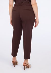 Hnědé kalhoty na míru s elastickým pásem, zužujícími se nohavicemi a dvěma zadními kapsami. Nosené s béžovými a hnědými sandály na podpatku.