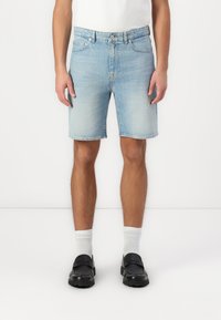 Shorts en denim bleu clair avec une coupe droite, cinq poches et une fermeture à bouton. Portés avec des chaussettes blanches et des chaussures noires.