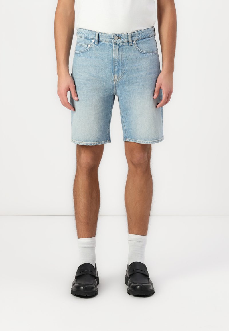Shorts en denim bleu clair avec une coupe droite, cinq poches et une fermeture à bouton. Portés avec des chaussettes blanches et des chaussures noires.