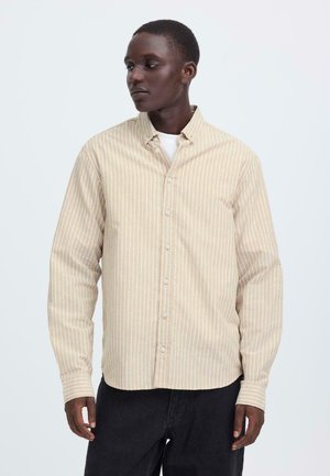 Beige Hemd mit weißen vertikalen Streifen, gefertigt aus weichem Stoff. Verfügt über einen Kragen und lange Ärmel, getragen über einem weißen T-Shirt.