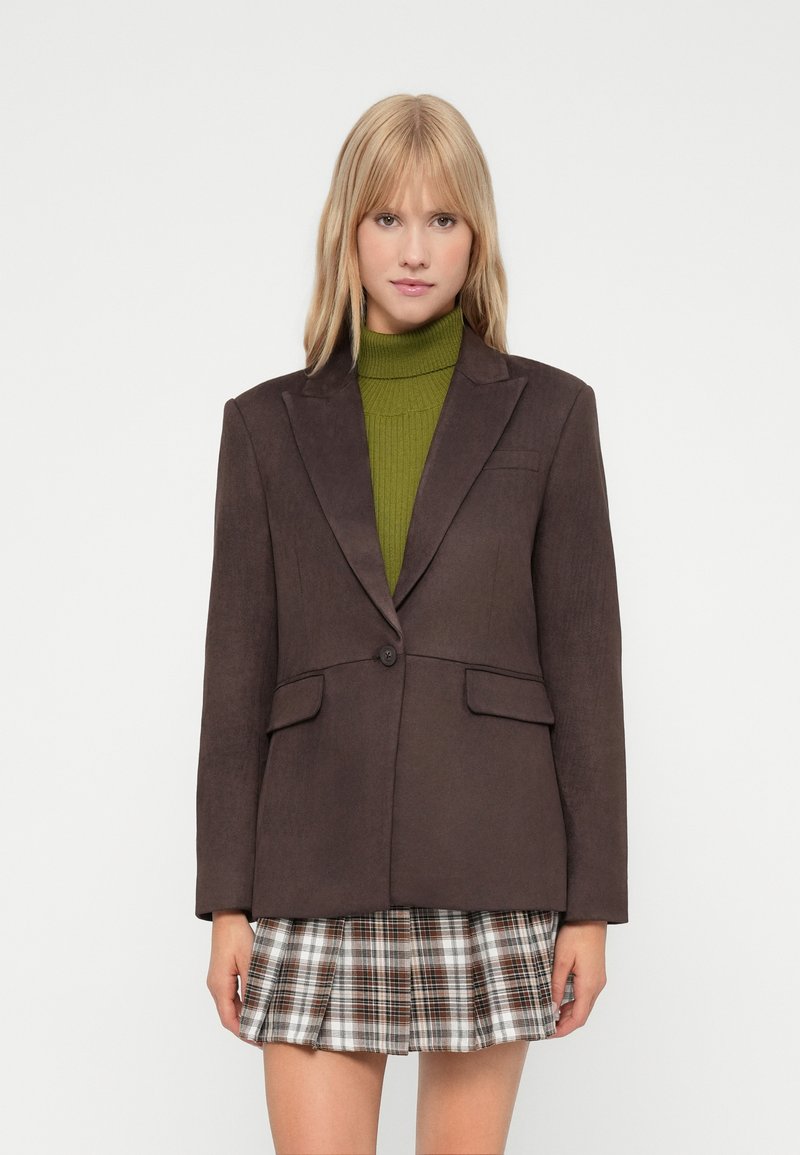 Bruine blazer met een notch lapel, enkele knoop, twee voorzakken en een gestructureerd ontwerp, gecombineerd met een groene coltrui en een geruite rok.