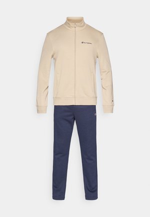 Beige jas met ritssluiting, hoge kraag en logo, gecombineerd met marineblauwe joggers. Beide gemaakt van zachte, rekbare stof.