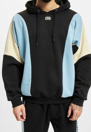 Mężczyzna ubrany w czarną bluzę z kapturem z niebieskimi i beżowymi zakrzywionymi panelami, sznurkami oraz dopasowane czarne spodnie z niebieskimi paskami po bokach.