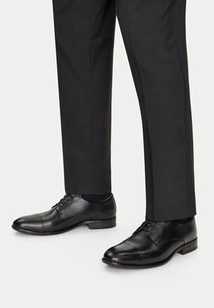 Juodos odinės suknelės bateliai su lygia apdaila, raišteliais ir apvaliu pirštu, derinami su tamsiomis pritaikytomis kelnėmis.