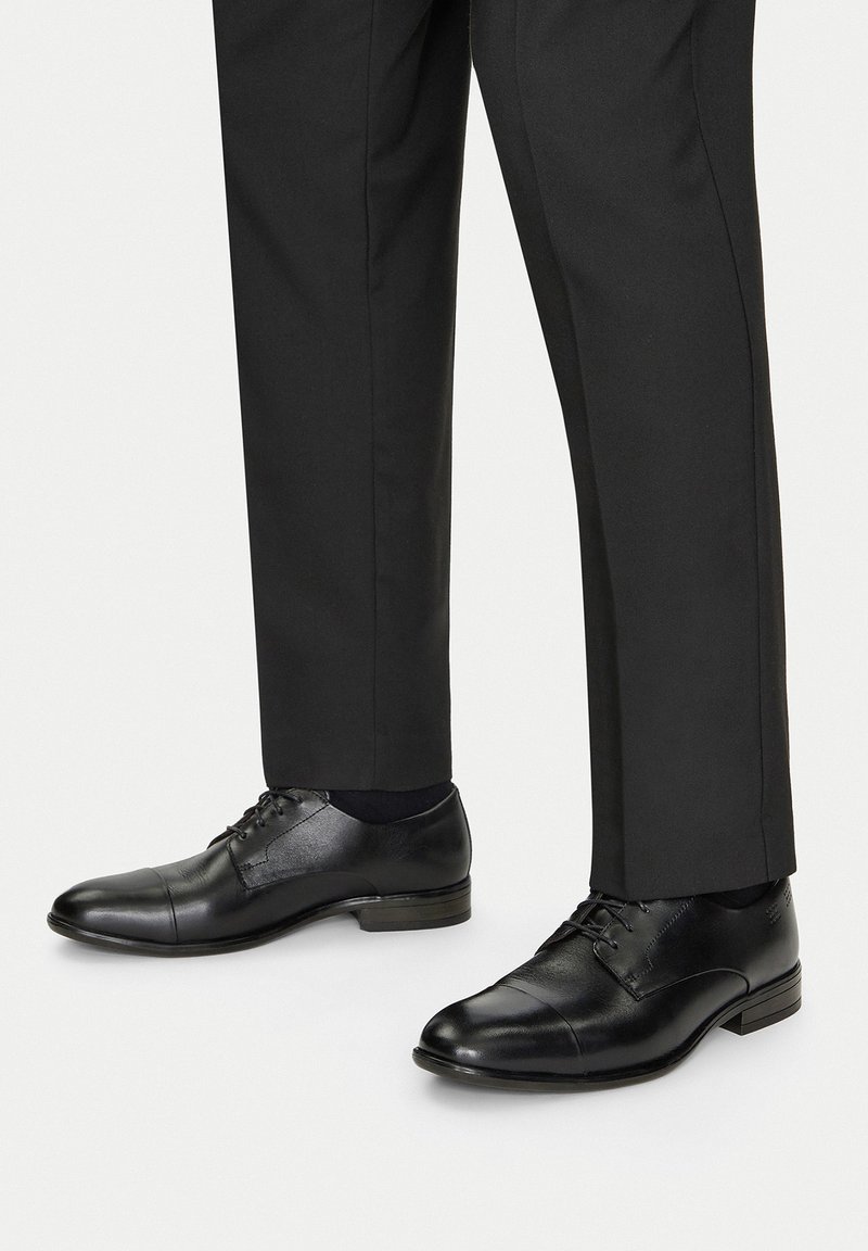 Mustad nahast kingad sileda pinnaga, nööbiga disainiga ja ümarate varvastega, mis on kombineeritud tumedate kohandatud pükstega.