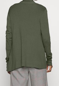 Olivgrön långärmad cardigan med avslappnad passform, ribbad textur och sidofickor, i kombination med svartvitt rutiga byxor.