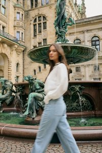 Femme en pull blanc et jean bleu marchant devant une fontaine en bronze verte ornée de statues dans une place historique européenne.