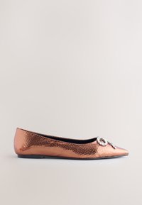 Chaussure plate en or rose avec une finition texturée et écailleuse. Bout pointu souligné par un ornement décoratif en cristal. Semelle plate.