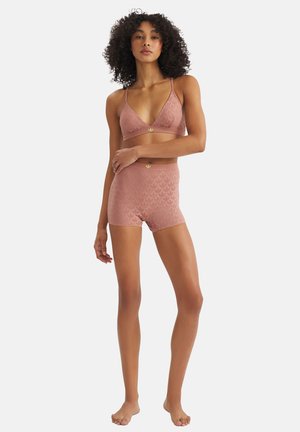 Mujer con cabello rizado, descalza, vestida con un conjunto a juego de bralette texturizado y pantalones cortos de cintura alta color rosa, sobre un fondo blanco.