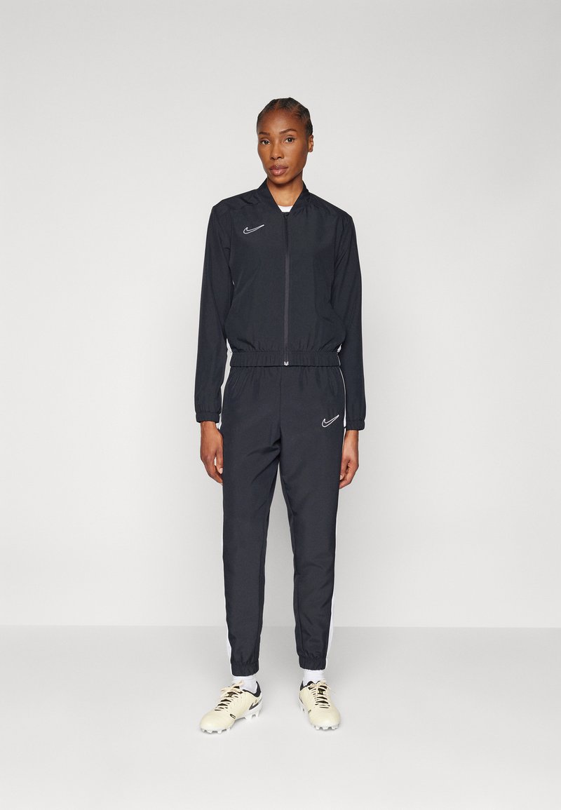 Giacca a vento nera con zip, polsini elasticizzati e pantaloni. Presenta il logo Nike bianco, tessuto leggero e una silhouette aderente.