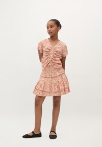 Een jurk met een bloemenpatroon, een geplooide voorkant, korte mouwen, gelaagde ruffles en een V-hals. Gecombineerd met zwarte platte schoenen.