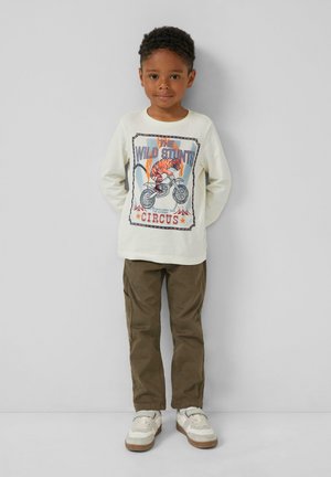Jongentje staat rechtop met zijn handen achter zijn rug, gekleed in een witte langemouwen shirt met een tijger- en motorfietsafbeelding, bruine broek en witte schoenen.