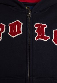 Hoodie zippé marine avec lettres brodées en relief blanches et rouges. Tissu lisse avec ourlet et poignets côtelés. Le matériel de la fermeture éclair est en argent.