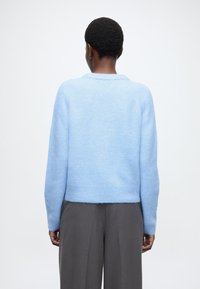 Světlomodrý krátký svetr s jemnou texturou a žebrovaným lemem, který má klasický límec a dlouhé rukávy, nošený se šedými kalhotami.