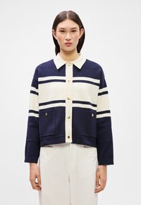 Tamsiai mėlynas ir kreminis dryžuotas megztinis su apykakle, auksiniais mygtukais ir priekinėmis kišenėmis. Pagamintas iš tekstūrinio nėrimo, trumpa dizaino ir ilgomis rankovėmis.