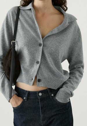 Femme portant un cardigan gris boutonné avec col, un jean taille haute foncé et un sac à main noir, les mains dans les poches du jean.
