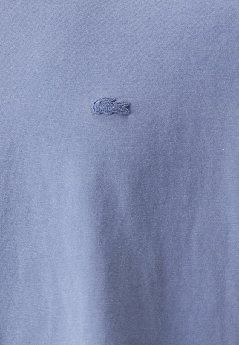 Camiseta de algodón azul claro con un logo de Lacoste bordado en azul oscuro con textura en el pecho. Tela suave y diseño sencillo con cuello redondo estándar.