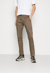 Pantalon en coton marron à coupe slim, avec poches avant et fermeture à boutons, associé à des chaussures de sport noires et grises.