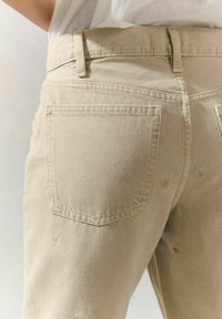 Jean en coton beige avec une coupe ajustée, un design classique à cinq poches et de subtils détails brodés sur les poches arrière.