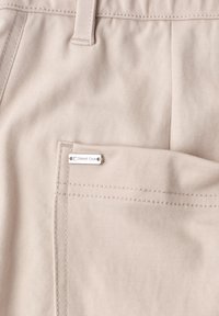 Pantalon en tissu beige en gros plan montrant la poche arrière avec une petite étiquette métallique de la marque « Street One » attachée.