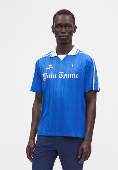 Homme portant un polo de tennis bleu avec col blanc, logos US Open et Polo Ralph Lauren, debout sur un fond blanc uni.