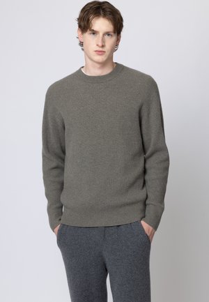 Giovane uomo dalla pelle chiara e capelli castani, indossa un maglione a coste verde oliva e pantaloni grigio scuro, in piedi con le mani nelle tasche.
