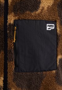 Chaqueta de forro polar marrón y marrón oscuro con un gran bolsillo rectangular negro con cremallera en el pecho y parche con el logo blanco de Puma.