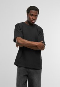 Zwarte korte mouwen t-shirt, ronde hals, relaxte pasvorm, lichte stof, gedragen met donkergrijze jeans, model staat met gekruiste armen.