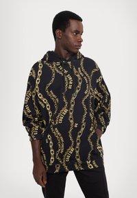 Musthave musta kapuutsiga, millel on kettmuster ja kuldne tekst "VERSACE JEANS COUTURE" kangal. Valmistatud pehmest materjalist, millel on mugav lõige.