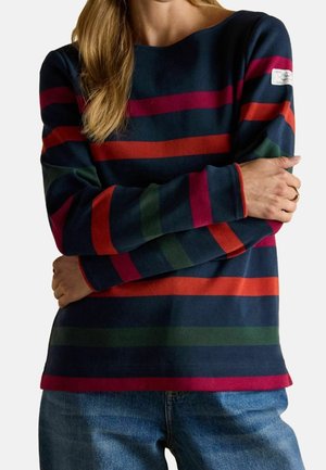 Personne portant un pull bleu marine à manches longues avec des rayures horizontales rouges, vertes et magenta, associé à un jean bleu et une bague en or.