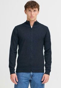 Sötétszürke, cipzáros pulóver magas nyakkal, kötött textúrával és hosszú ujjakkal, kék farmerrel párosítva. Egyszerű és letisztult dizájn.