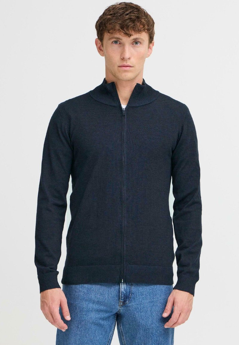 Sötétszürke, cipzáros pulóver magas nyakkal, kötött textúrával és hosszú ujjakkal, kék farmerrel párosítva. Egyszerű és letisztult dizájn.